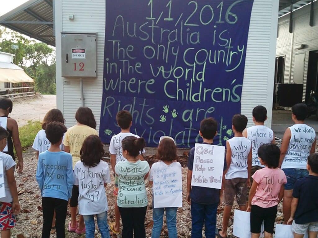 A group of children with their backs turned to the camera. They are wearing t-shirts with writing on the back. They stand in front of a building with a large sign.