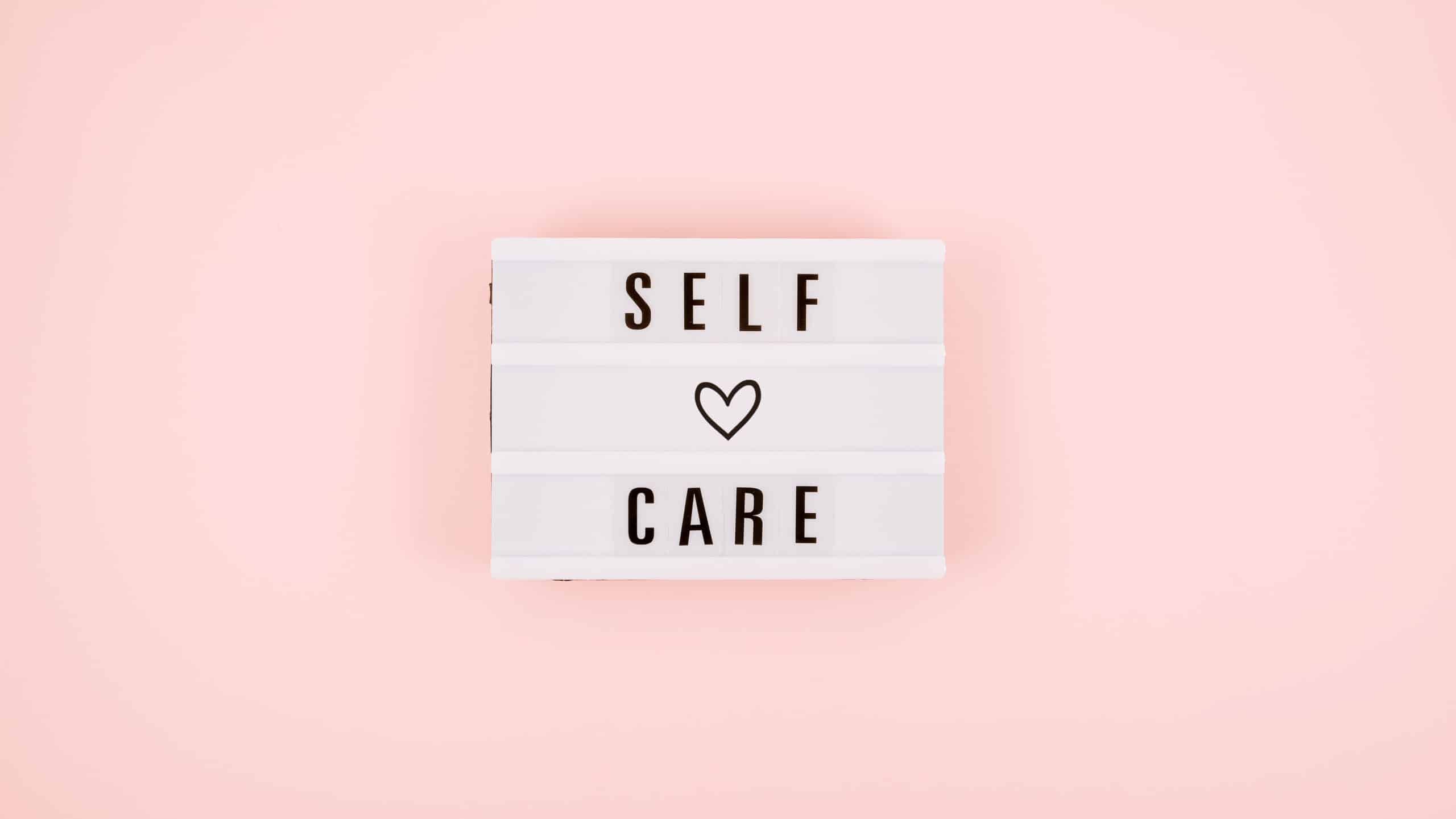 A white sign with black words spelling "self care" with a black heart. On a pink background.