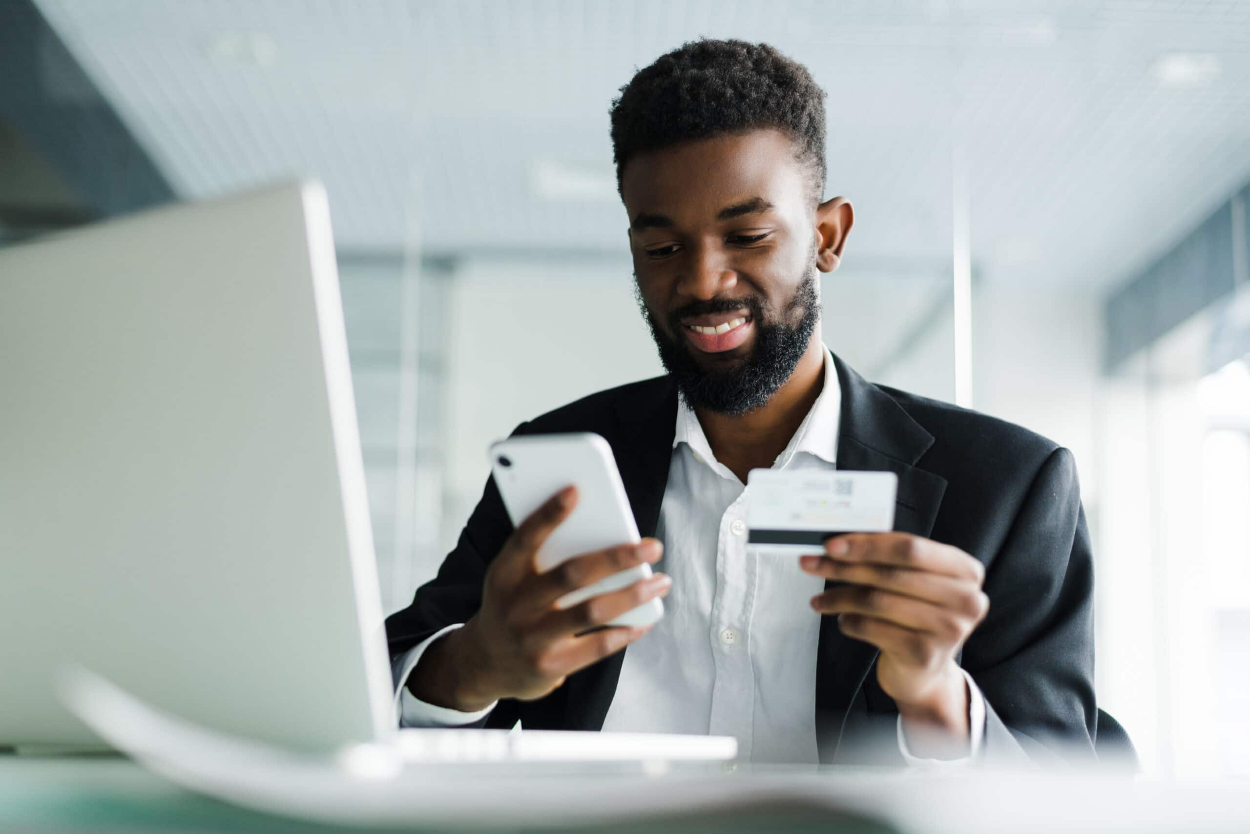 An African American man sits at a laptop computer. He holds a phone in one hand and a credit card in the other hand.