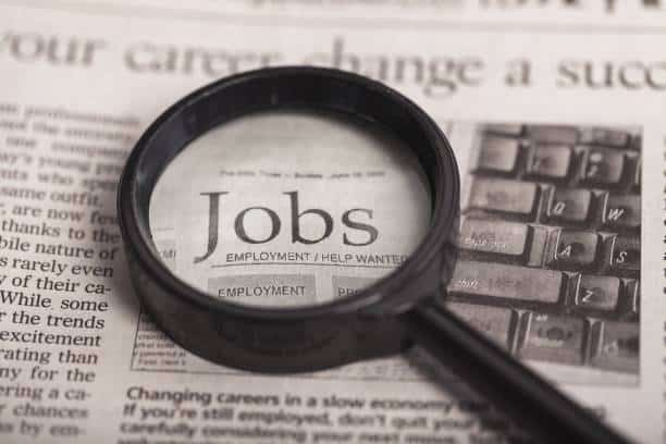A black magnifying glass sits on top of a newspaper. The word "Jobs" is highlighted under the magnifying glass.