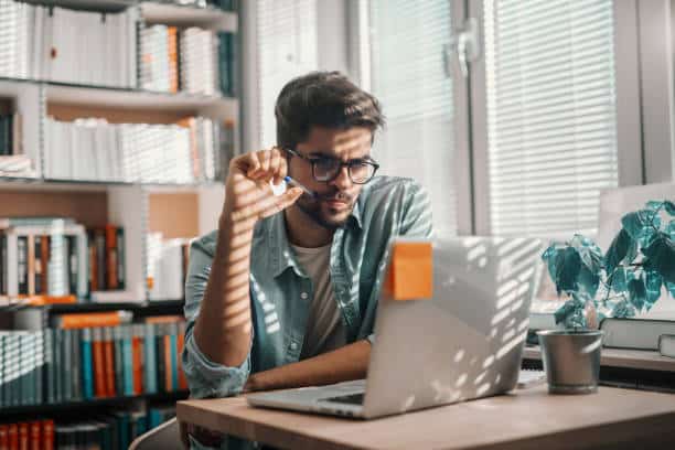 A man sits at a desk in front of a computer. He is looking at the computer and holds a pen in his hand.