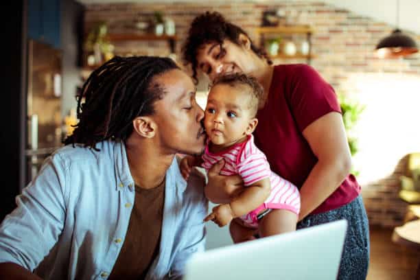 A man is turned to the side and kisses a baby that is held by a woman. She is smiling and looking at the baby.