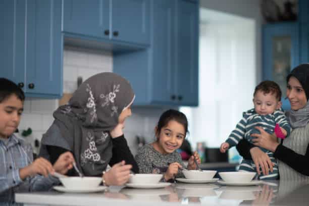 A woman in a hijab sits with several children at a table. In front of them are bowls.