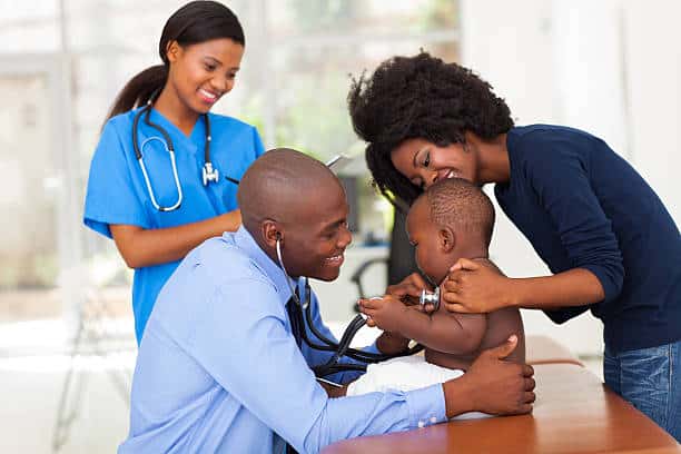 A doctor examines a baby with a stethescope. A woman holds the baby's shoulders. A woman wearing a blue uniform and aa stethescope stands to the side.