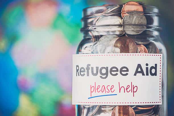 Glass jar full of coins against a multi-colored background. There is a sign on the jar that reads, "Refugee Aid please help"