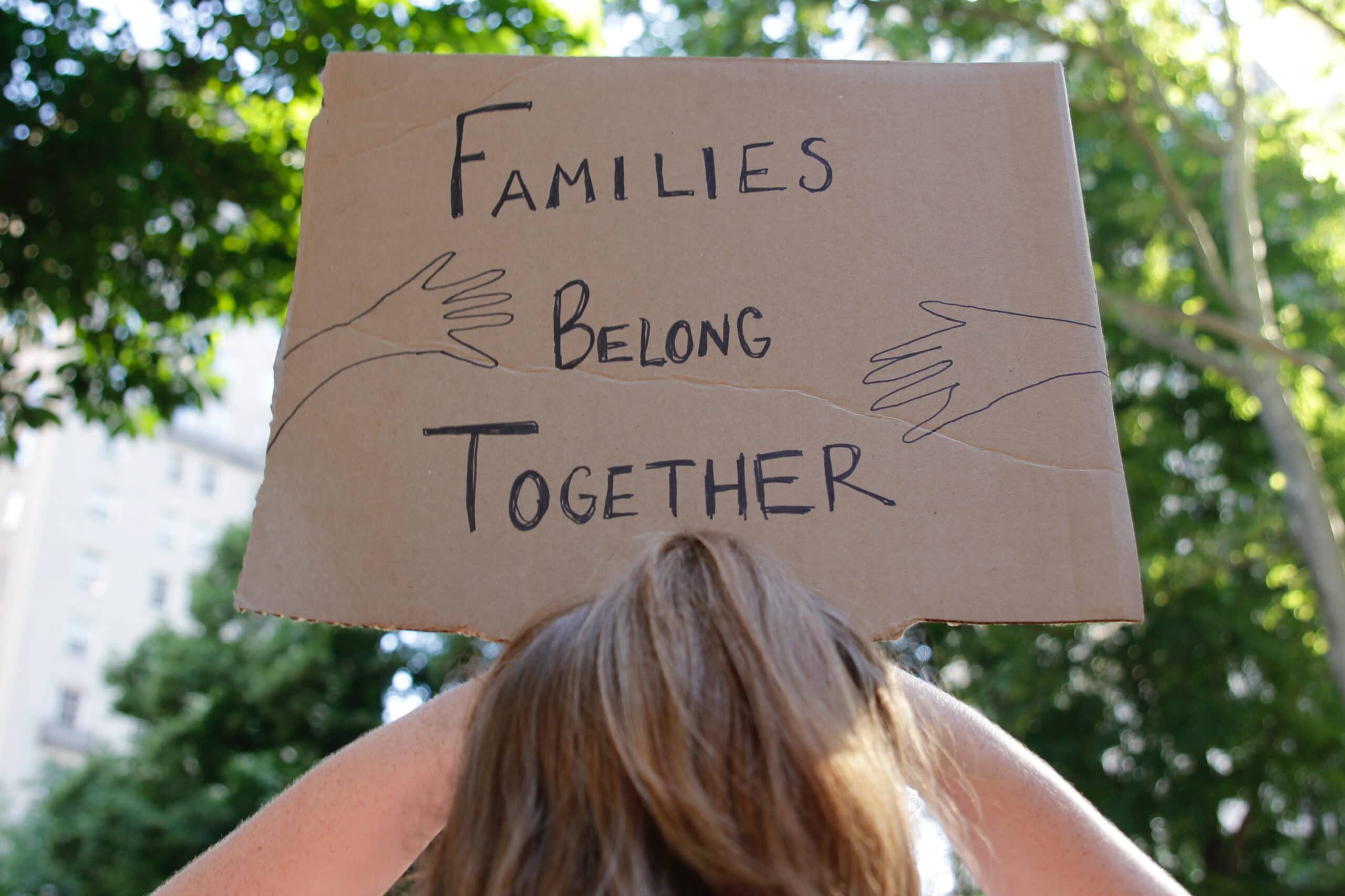Two hands hold aa cardboard sign with the words "Families belong together" written on it in black.