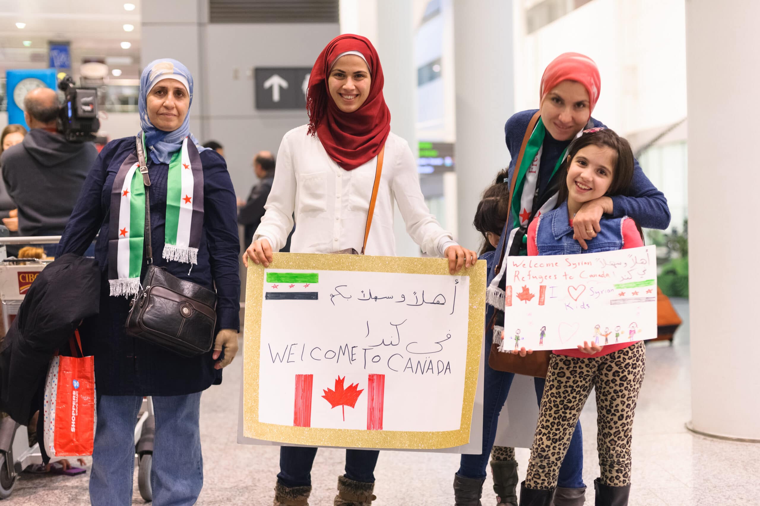 A group of people face the camera. A woman in a hijab holds a sign that says "welcome to Canada". A child holds a welcome sign. They are surrounded by two other people.