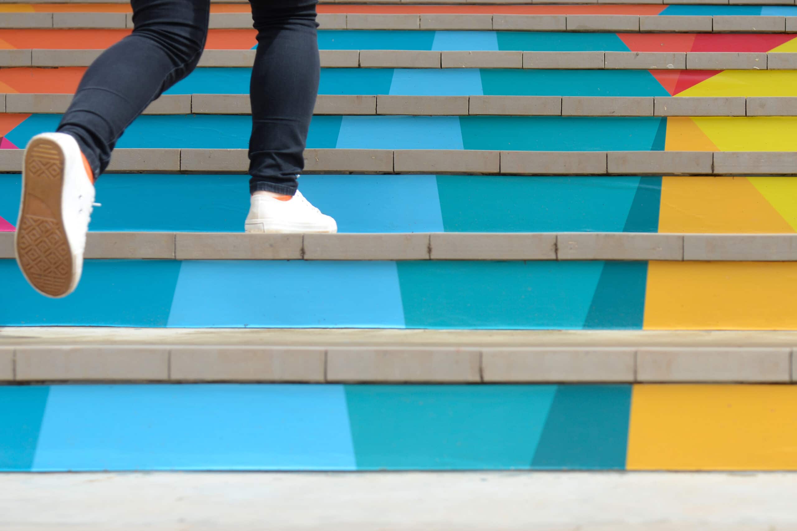 Lower part of legs wearing jeans and white tennis shoes, walking up multi-colored stairs.