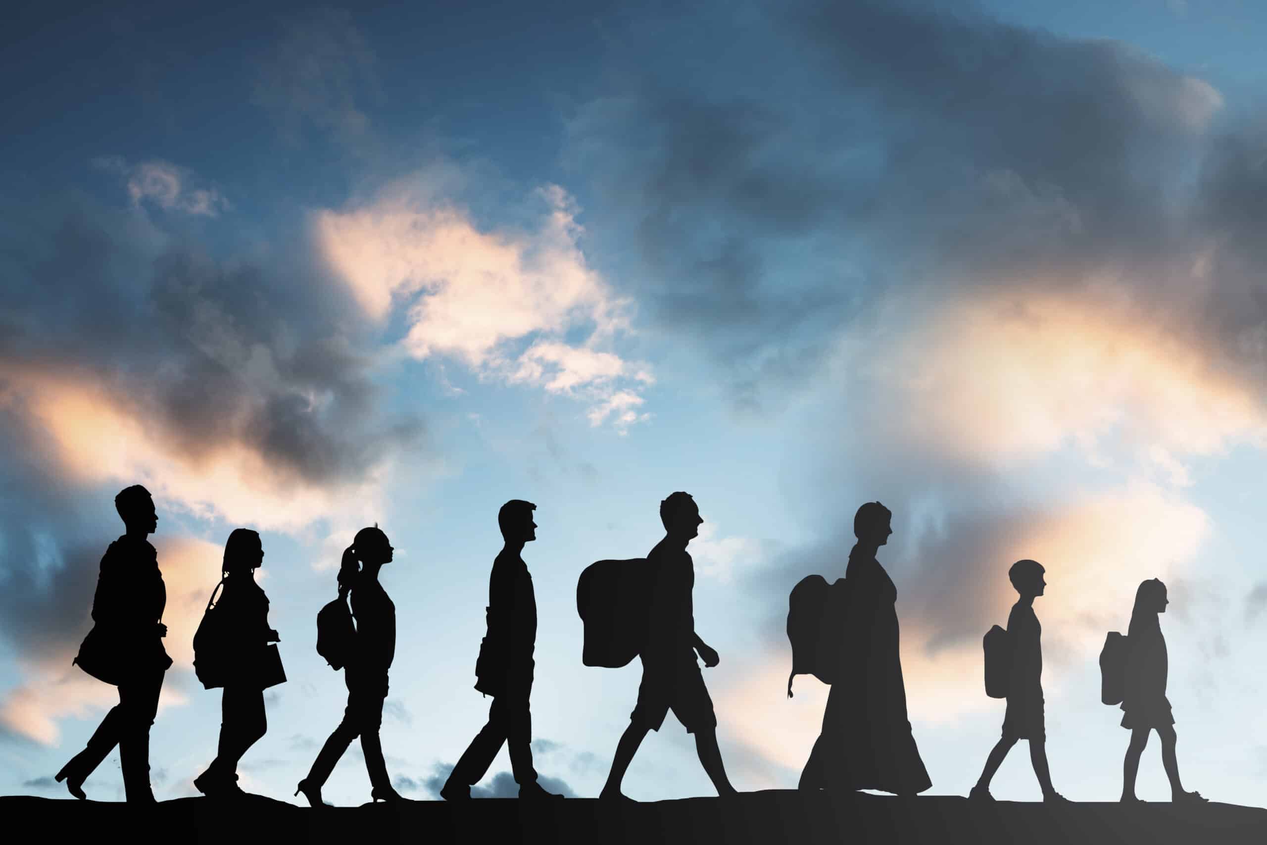 A group of silhouetted figures walks across the ground. The sky is blue and there are dark clouds. Some of the figures are carrying suitcases.